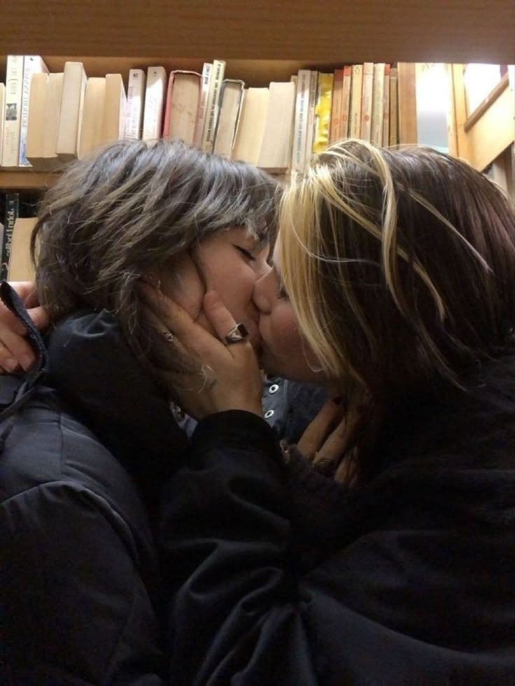 two women are kissing in front of a bookshelf filled with books. they are both wearing jackets and one has her hand on the other's face. the woman's hand has a ring on it. they are standing very close to each other.