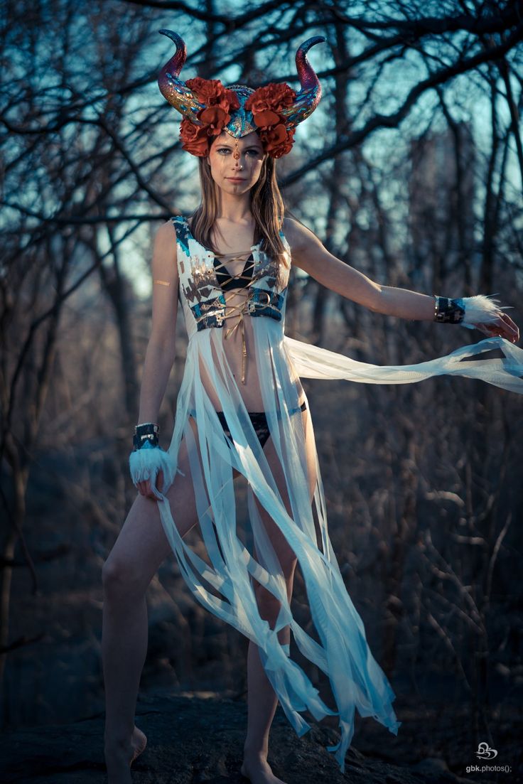a woman stands confidently in a dimly lit forest, her strong pose accentuated by her attire. she wears an elaborate horned headdress adorned with red roses, a whitelaced top and a flowing white skirt.  her attire, coupled with her determined expression, suggests a warrior queen or a mythical forest deity.