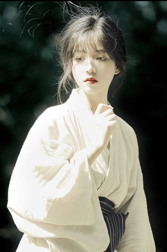 a young woman with dark hair and red lipstick is wearing a white kimono. she is standing outdoors in soft, natural light and looking down with a serene expression. the background is a blur of green.