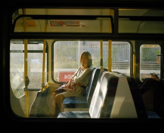 a woman sits alone on a bus, bathed in sunlight streaming through the window. she appears calm and pensive, looking out the window. the interior of the bus is visible, showing seats and yellow handrails.  the overall mood is peaceful and contemplative.