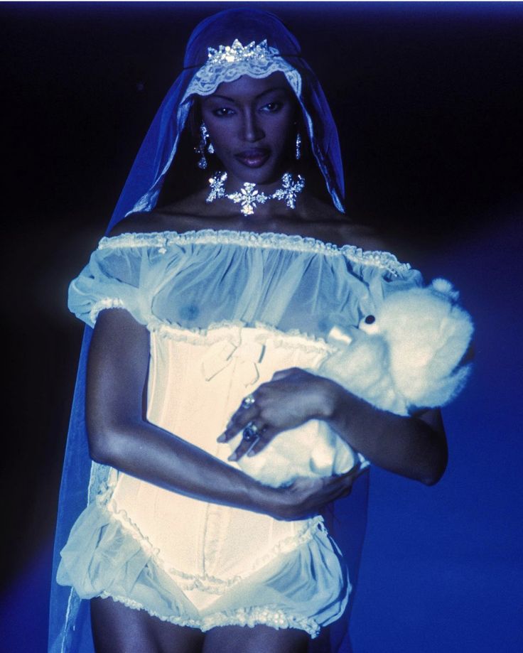 the image is a striking photograph of supermodel naomi campbell on a fashion runway. she's dressed in a bridalinspired ensemble that incorporates elements of lingerie. the outfit consists of a sheer, offtheshoulder top, a white corset, and a ruffled, lingeriestyle bottom.  she wears a delicate tiara or crown and a long veil, contrasting with the more risqué corset.  a snowflakeshaped necklace and earrings complete the look.  she is holding a white plush toy, adding an unexpected softness to the overall edgy, highfashion presentation. the overall palette is dominated by shades of blue, lending a cool and somewhat ethereal quality to the image.  the photograph is likely a vintage shot from a 90s fashion show, capturing the era's unique blend of high fashion and sometimes provocative aesthetics.