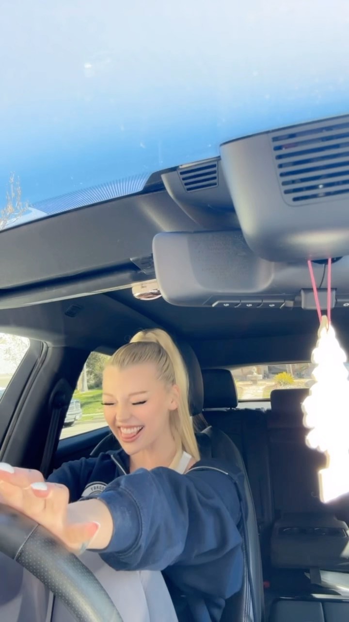 a woman with blonde hair pulled back in a ponytail is smiling and laughing while sitting in the driver's seat of a car. she is wearing a navy blue sweatshirt and her hands are on the steering wheel. above her is a sunroof. a white, fluffy car air freshener hangs from the rearview mirror. the car interior is dark, and the lighting is bright, indicating a sunny day.