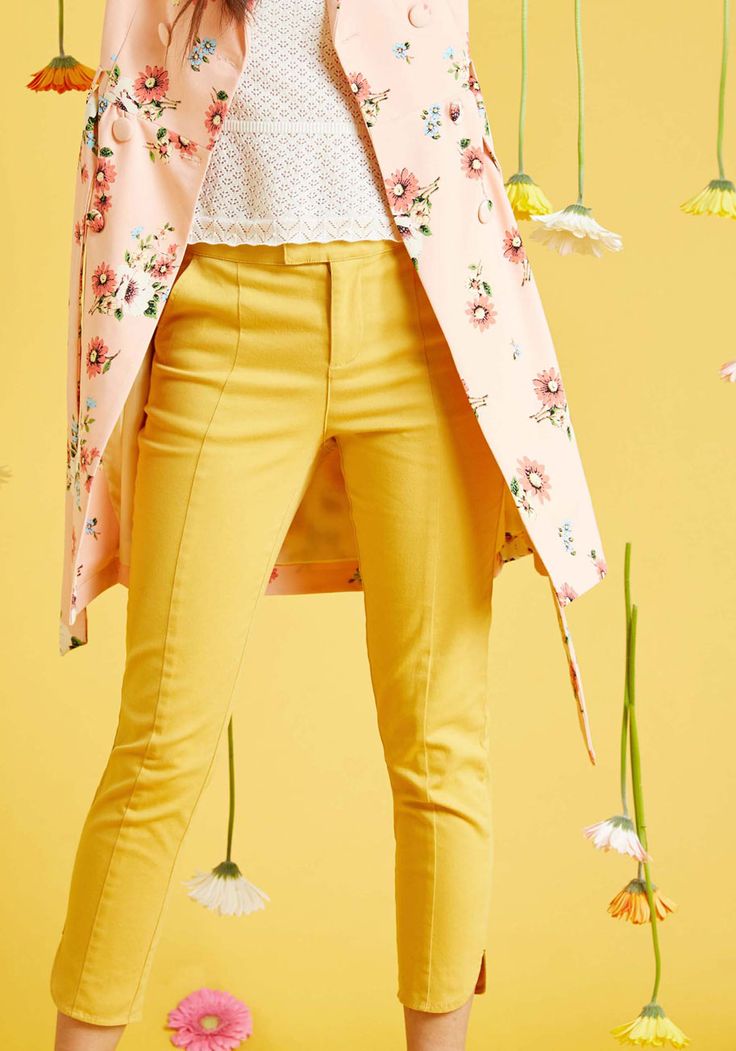 a woman is wearing a pink floral jacket and bright yellow pants, a white lace top. she stands in front of a yellow background with various flowers.