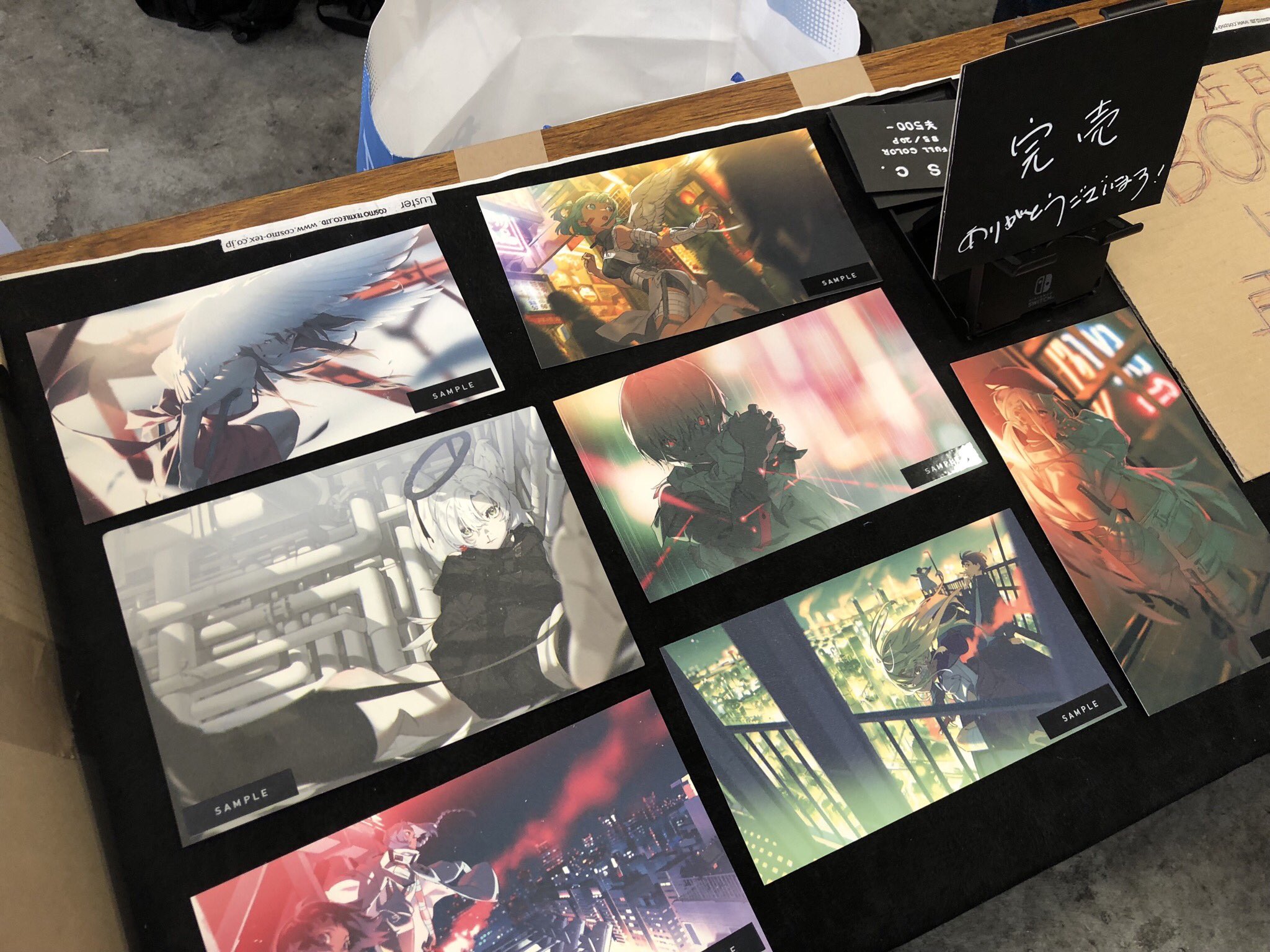 the image shows a table topped with anime art prints at what appears to be an anime convention. each print is marked with "sample" and leans against a larger sign. the sign  indicates the prints were being sold for ¥500 and are now "sold out", with a message thanking customers. the overall color scheme is dark with pops of color from the artwork.