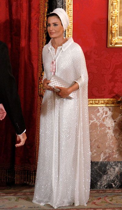a full shot of princess lalla salma of morocco, standing in what appears to be a palace room with red walls and goldframed artwork.  she is wearing a long, elegant white sequined abaya, a traditional moroccan garment, and a white hijab covering her head. she's holding a white clutch. a portion of a person in a dark suit is visible on the left side of the frame. the overall atmosphere conveys luxury and royalty.