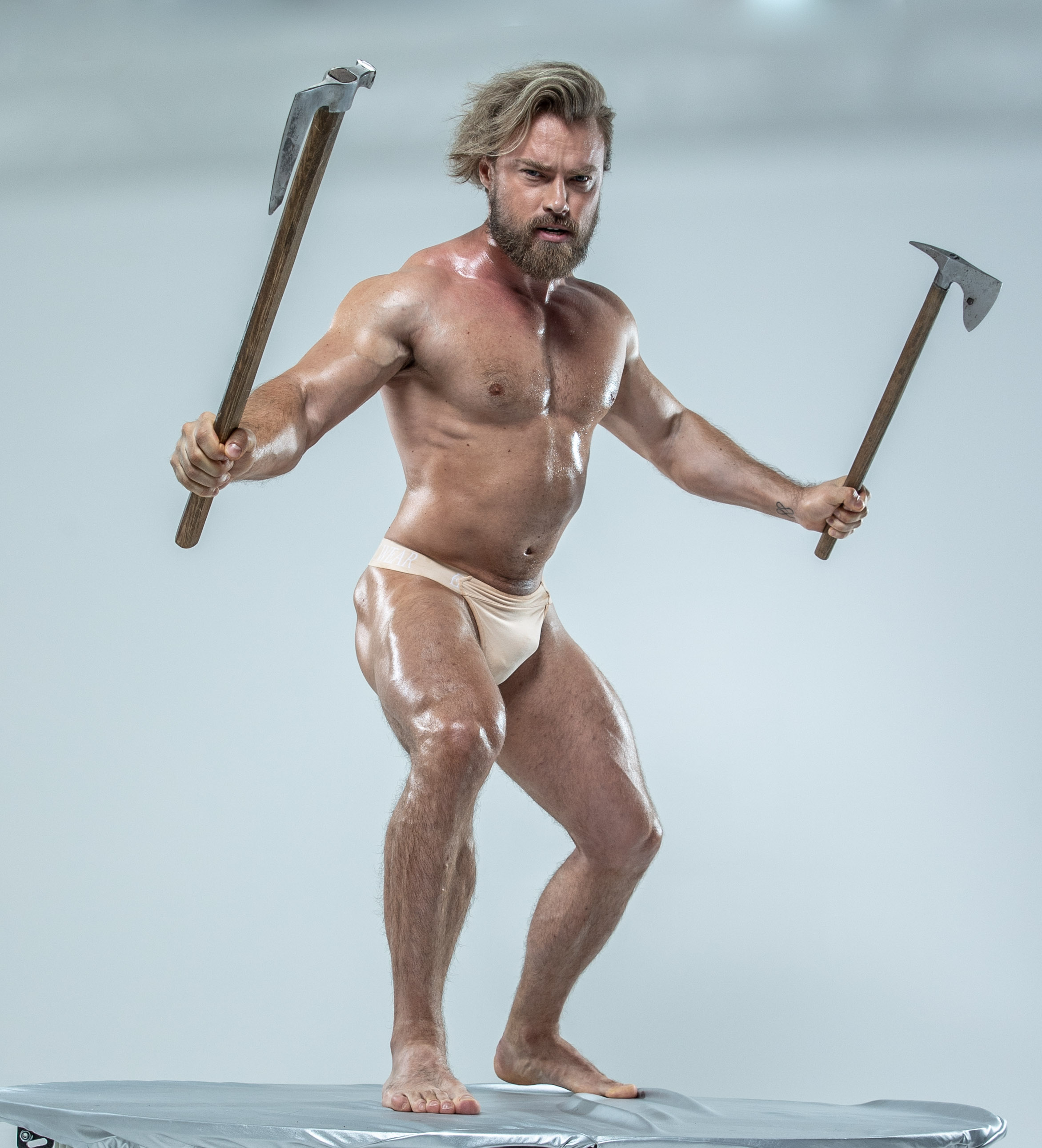muscular, bearded man stands on a platform against a white backdrop. he is holding two axes, one in each hand, in a powerful pose. his expression is intense and determined. the lighting is bright and artificial, highlighting his physique. the overall image conveys strength, power, and a warriorlike presence.