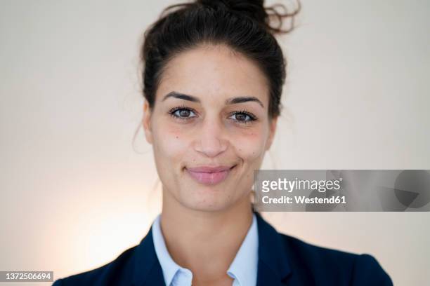 the image is a portrait of a woman with olive skin and dark hair pulled up in a bun. she is wearing a navy blue blazer over a white collared shirt and is smiling softly at the viewer. the background is a light beige color and the image appears to have been taken in a studio setting with professional lighting.