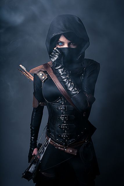 this is a portrait of a woman dressed as a warrior. she is wearing a dark hooded costume with a leather bodice. she is holding a bow and arrow, and her face is hidden by a mask. the background is dark and smoky, giving the photo a mysterious and dangerous feel. she is standing in a powerful and confident pose, radiating strength.