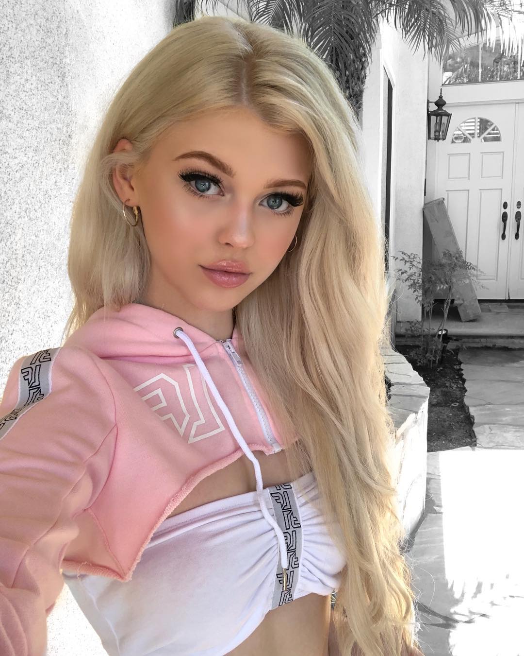 a closeup shot of loren gray, a young woman with blonde hair and blue eyes, wearing a pink cropped hoodie over a white top. she is looking directly at the camera with a slight smile. she is standing outdoors, with a palm tree and the facade of a house visible in the background. the lighting is bright and flattering, highlighting her features and creating a soft, ethereal atmosphere.