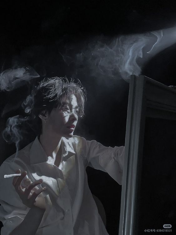 a man in a white shirt is illuminated by a beam of light in a dark room. he is holding a cigarette in his left hand and a paint brush in his right hand. there is an easel with a canvas on it to his right and smoke from his cigarette billowing around his head.