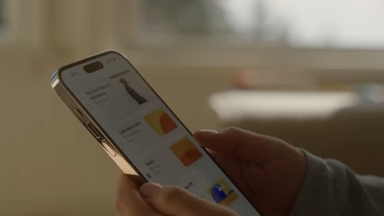 a closeup shot of a person holding a smartphone with a gold frame.  the screen displays a vertically scrolling feed with various app icons and titles. the background is blurred, suggesting a home environment with soft, natural lighting. the hand holding the phone partially obscures the lower portion of the screen.