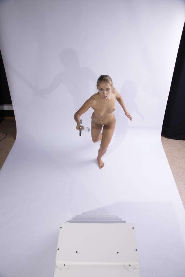 a nude woman is running towards the viewer with a determined expression. she is holding an object in her hand, possibly a tool or weapon. the background is a plain white studio backdrop. the image is a full shot taken from a high angle, emphasizing the subject's forward motion and the sense of urgency.