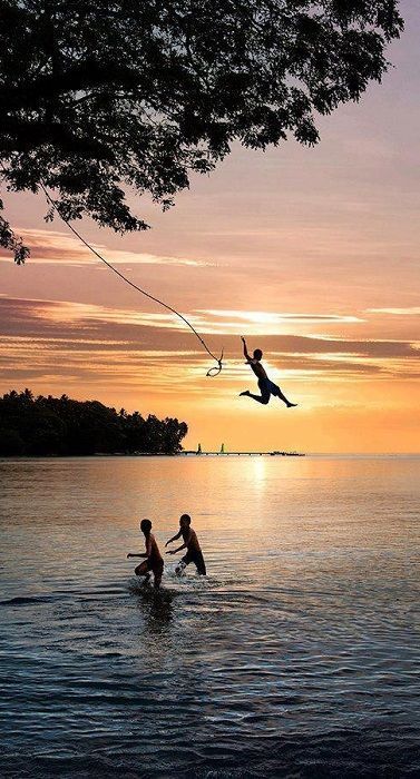 a silhouette of a young boy swinging on a rope swing over the ocean at sunset.  two other boys are in the water below. the sky is a beautiful orange and purple with some white clouds.