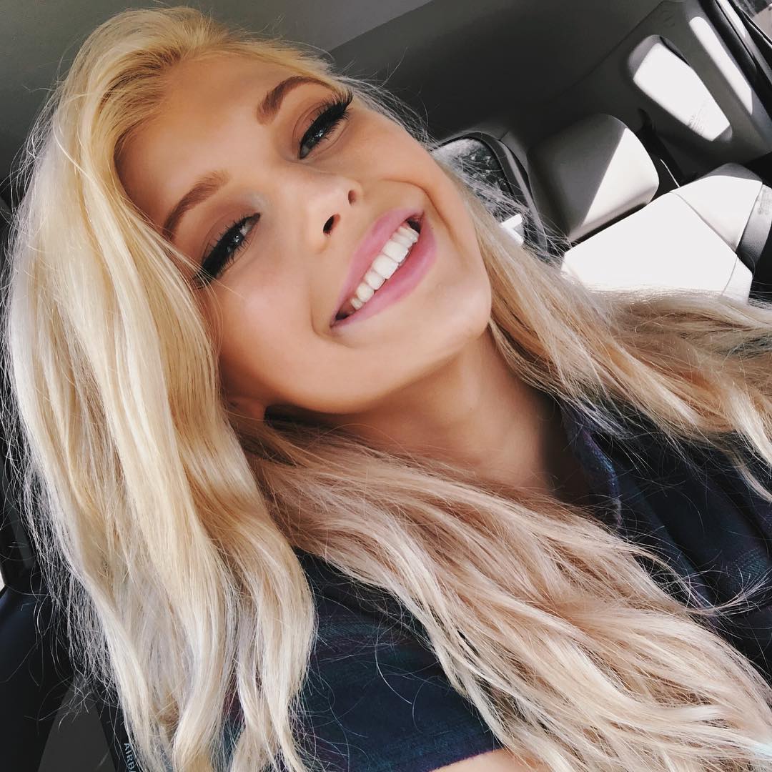 a young woman with long blonde hair is smiling at the camera in a selfie. she has on makeup including eyeliner and mascara. she appears to be inside a car, wearing a plaid shirt. her teeth are visible in her smile.