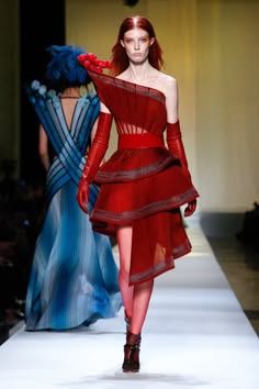 two female models wearing stylish outfits walk down the runway. the model in the foreground is wearing a bright red dress and heels. the model in the background is wearing a blue dress and heels.  both models have their hair styled in a unique manner and look elegant and stylish.