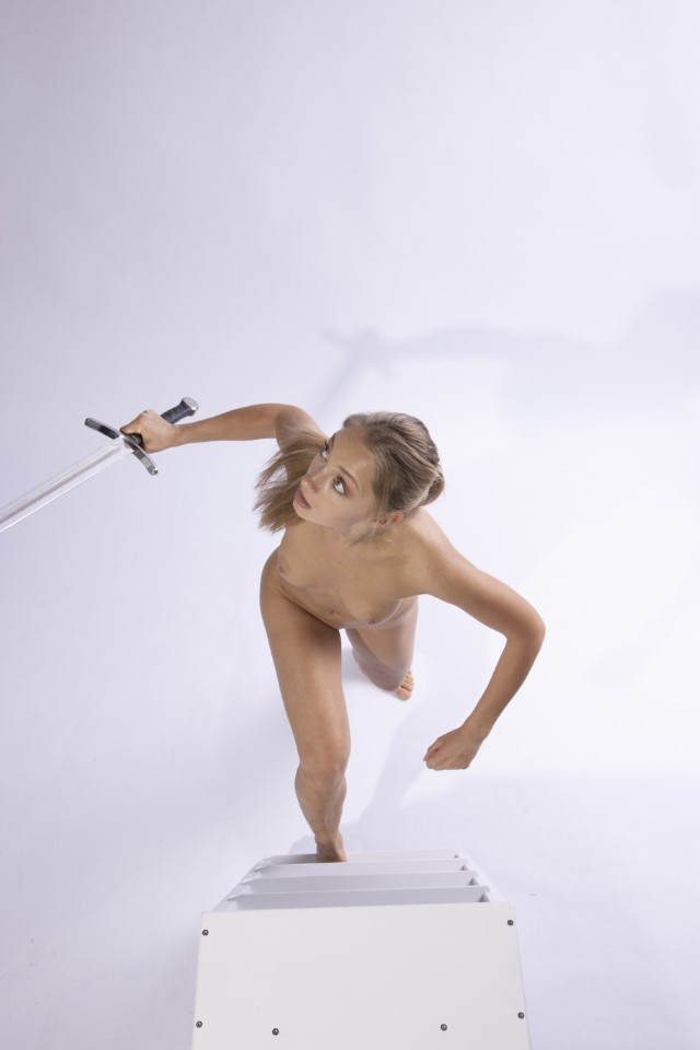 a highangle studio shot depicts a nude, fairskinned woman with blonde hair, holding a sword in her right hand, looking up towards the camera. she is standing on a set of stairs. the background is plain white.