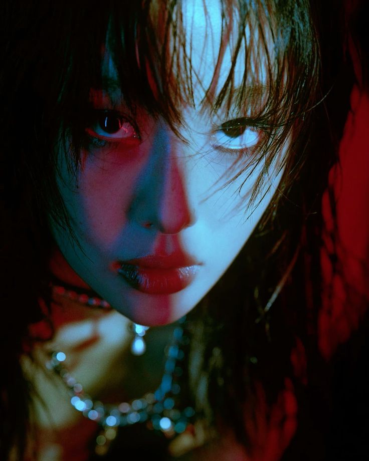 a closeup portrait of a woman's face. the lighting is dramatic, with red light coming from one side and blue light illuminating the front of her face. her hair is dark and appears wet or slicked back. she is wearing a necklace, and her expression is intense. the overall mood is dark and mysterious.