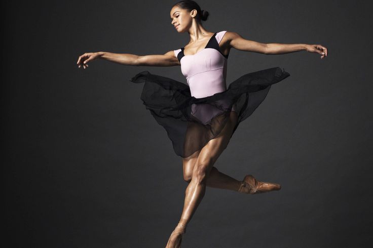 the image captures the captivating performance of misty copeland, a renowned ballerina, gracefully frozen midair during a ballet leap. set against a minimalist gray backdrop, the studio lighting accentuates her powerful physique and the flowing lines of her black dance skirt. her focused expression and extended limbs speak volumes about the dedication and artistry inherent in ballet.
