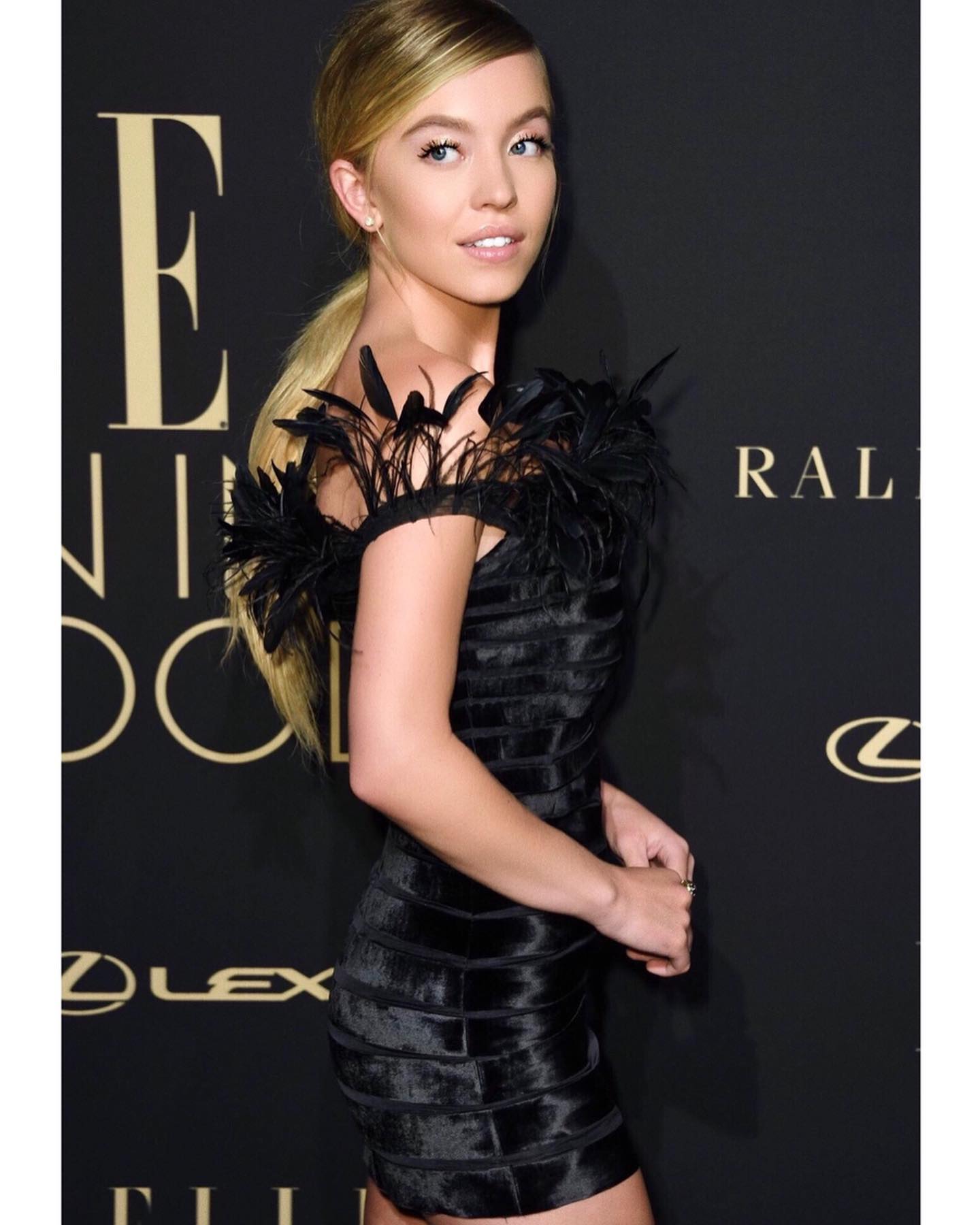 the image shows a young blonde woman, possibly a celebrity,  wearing a stylish black mini dress adorned with feather trim at the shoulders. she has her hair styled in a ponytail and is posed in a side profile, looking over her shoulder. the backdrop suggests she is at a formal event or red carpet, with visible logos for elle women in hollywood, ralph lauren, and lexus in the background. the overall impression is one of elegance and glamour.