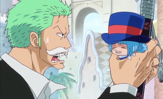 zoro is yelling with a vein bulging out of his head while holding a miniature chopper by the hat. chopper has a pained expression on his face as zoro holds him aloft.  they appear to be in a courtyard of some sort, with white stone buildings visible behind them.