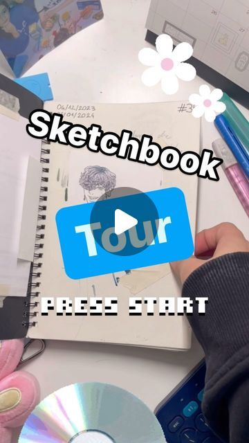 he image shows a sketchbook with an animestyle character drawing. on top of the sketchbook, text overlays read "sketchbook tour" and "press start," suggesting a video tutorial or walkthrough of the artist's sketchbook.  various art supplies, including pens and a calculator, are scattered around the sketchbook on a desk.  the overall aesthetic is pastel and incorporates decorative elements like daisies. the video play button graphic further emphasizes the video content.