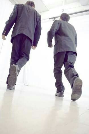 two men in suits are walking away from the camera down a white hallway. the perspective is from a low angle, looking up at the men. the lighting is bright, with natural light coming from overhead. the overall feeling is one of business and purpose.