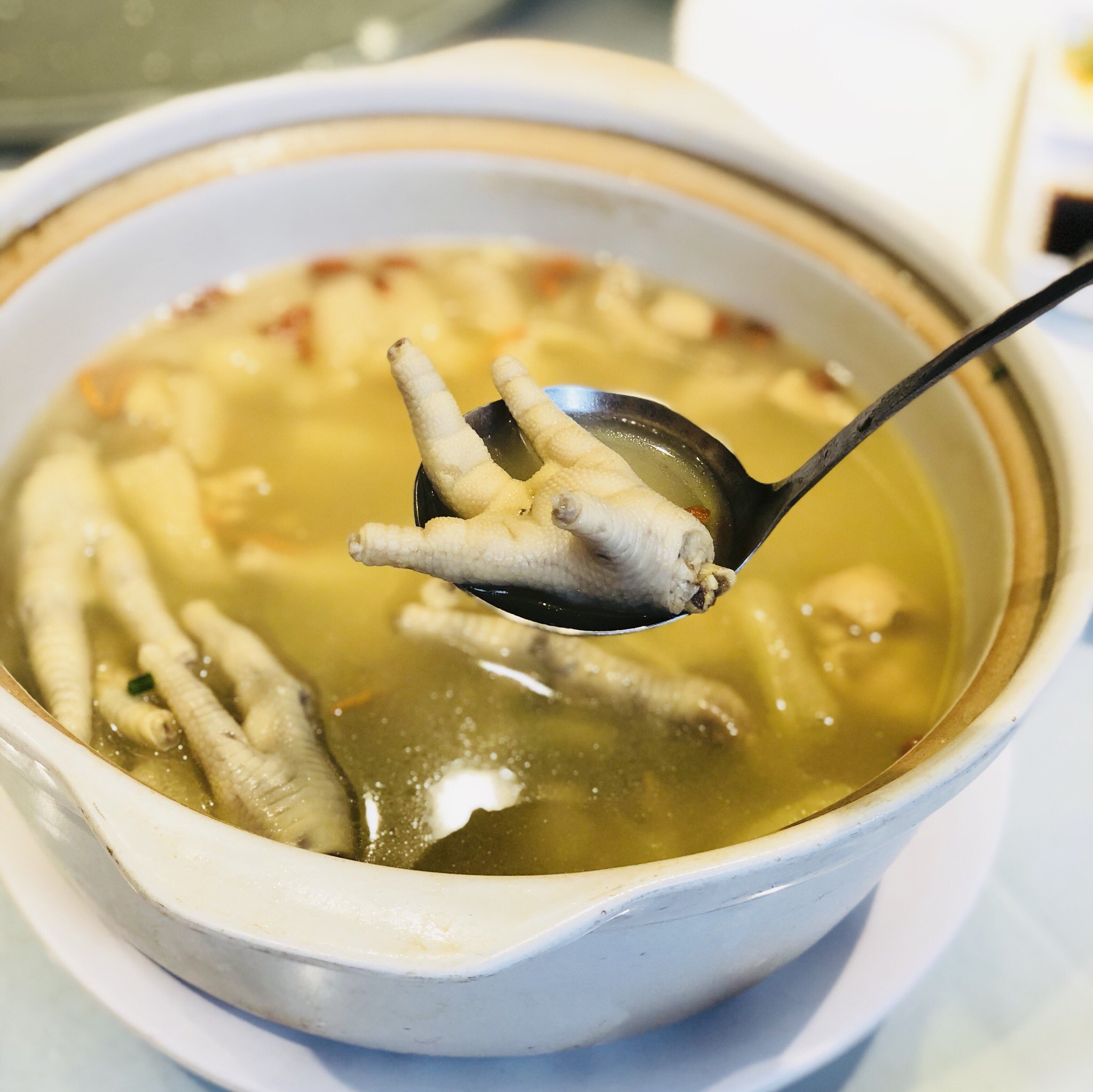 closeup view of a bowl of steaming hot chicken feet soup, a popular asian dish. the soup has a clear yellowish broth and contains chicken feet, herbs, and other ingredients. the image is taken indoors in a restaurant setting.
