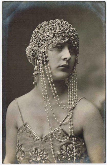 a black and white portrait of a young woman with a beautifully beaded headpiece and necklace, she is wearing a lowcut top and a soft expression on her face. her jewelry and her pose give her an elegant and glamorous look.  the photograph was taken in the 1920s, and its style evokes the fashions of the time, with a touch of art deco and flapper style.