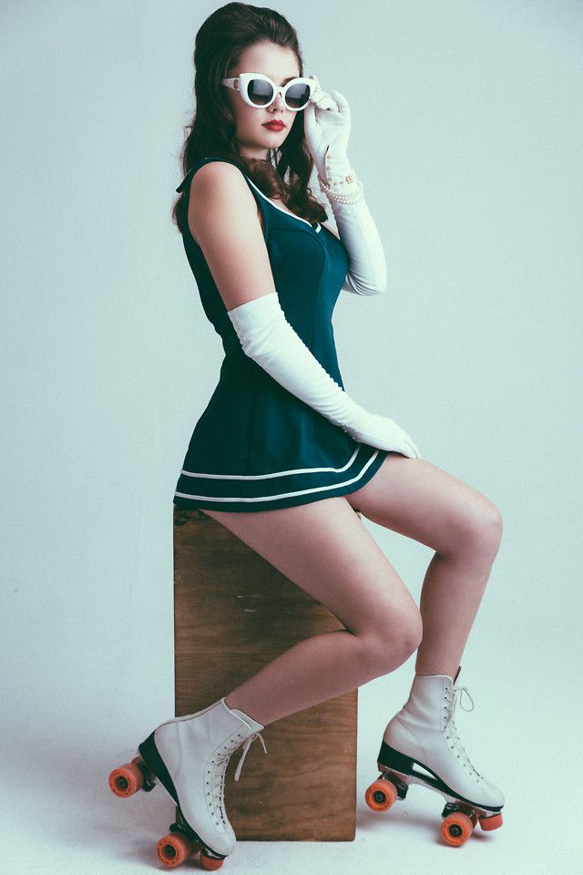 a woman with a vintage aesthetic, possibly inspired by 1950s or 1960s fashion, poses in a studio setting.  she is wearing a navy blue, sailorstyle romper, white roller skates, white gloves, and white cateye sunglasses. she is seated on a simple wooden block,  accentuating the retro feel of the image.  her pose and attire suggest confidence and a playful attitude.