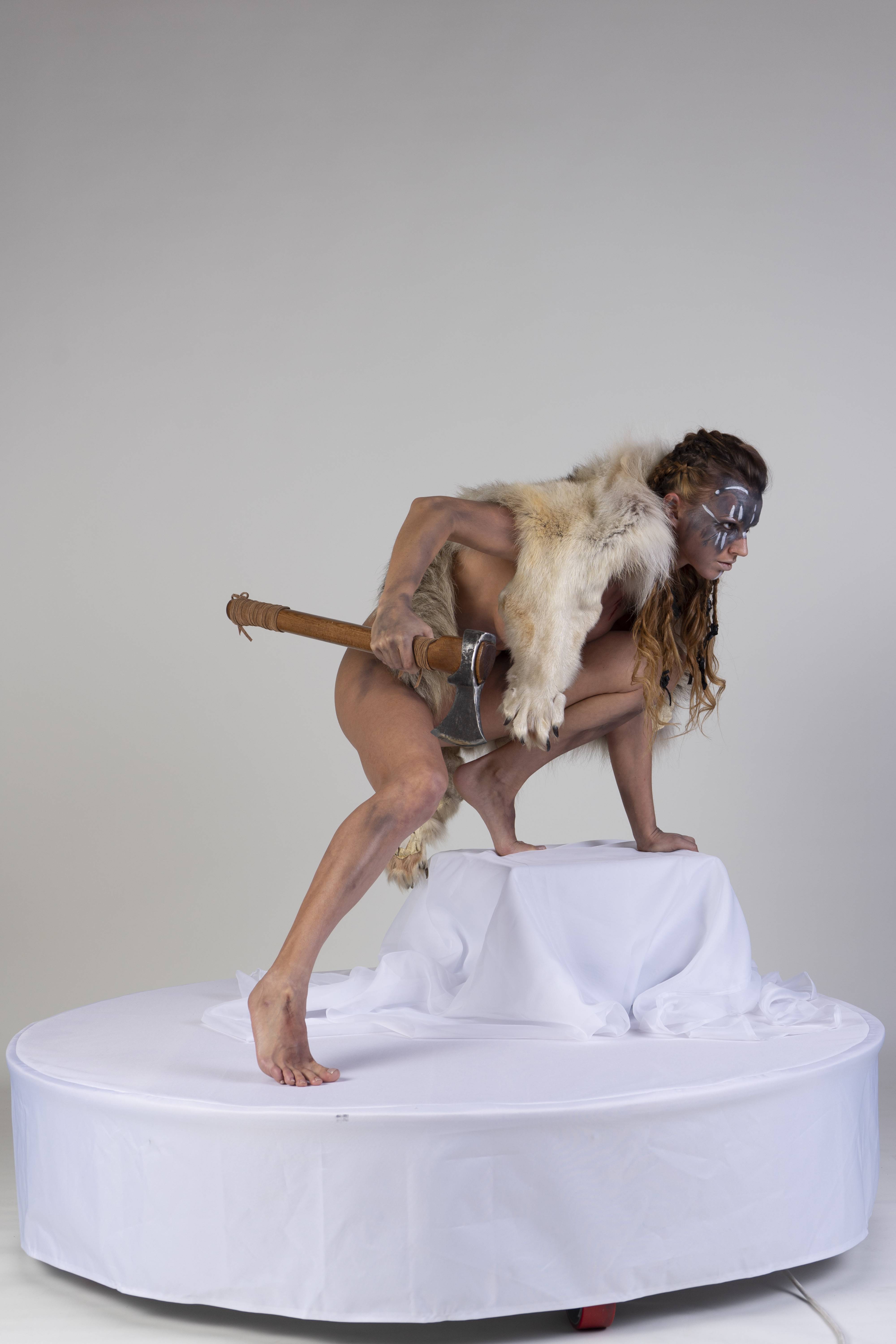 a woman dressed as a tribal warrior is crouching on a white surface. she is wearing a fur headdress and has makeup on her face. she is holding an axe in her right hand. the background is a plain white studio shot.