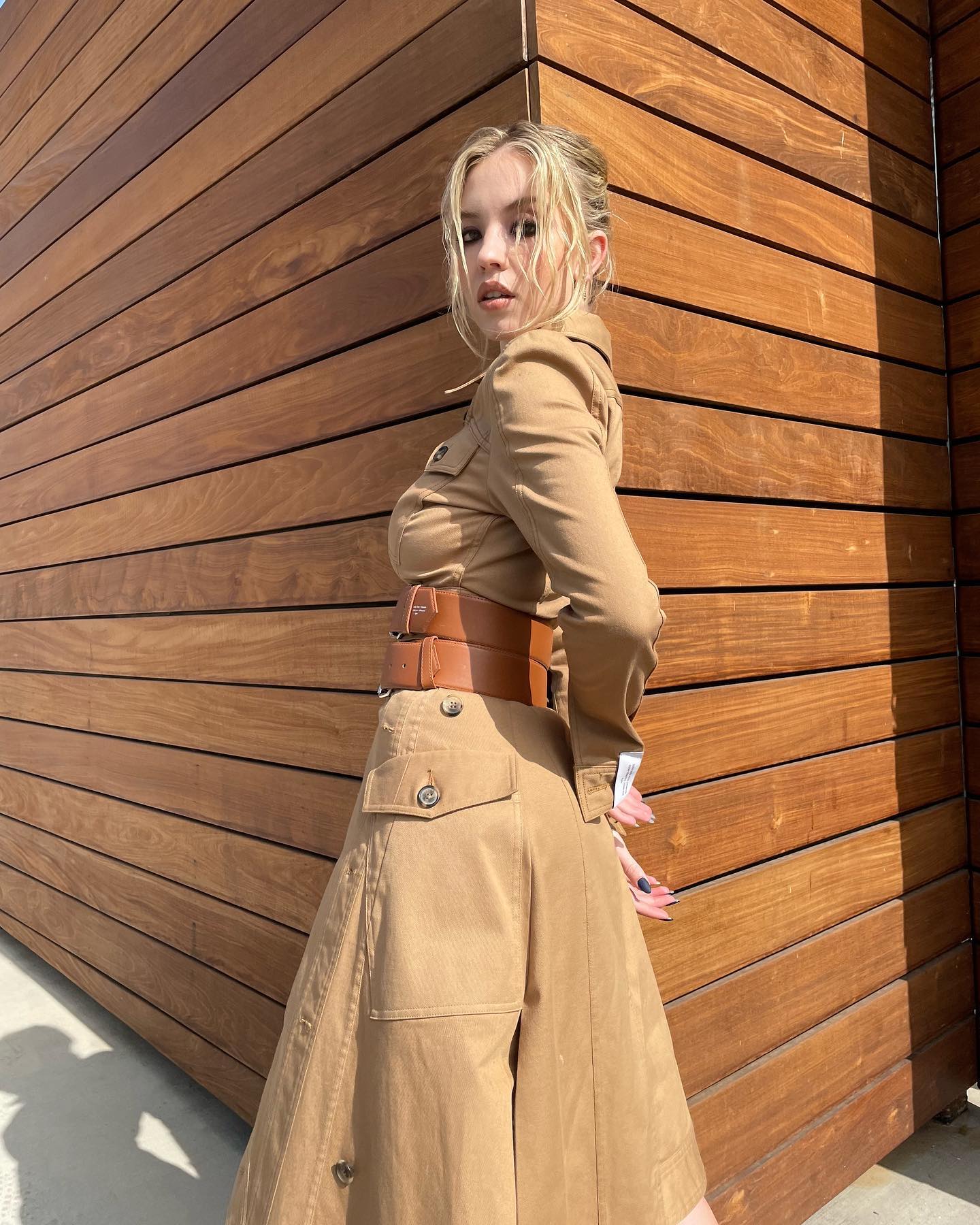 a blonde woman is standing outdoors, angled slightly away from the camera, posing against a wall made of horizontal, brown wooden planks. she's wearing a stylish, tancolored, longsleeved shirtdress that cinches at the waist with a wide brown leather belt. the skirt of the dress flares out below the belt. the dress has multiple pockets and button detailing. the overall style is casual yet chic. the sunlight casts shadows on her and the wood. the scene is bright and welllit, giving a summery or fall vibe.