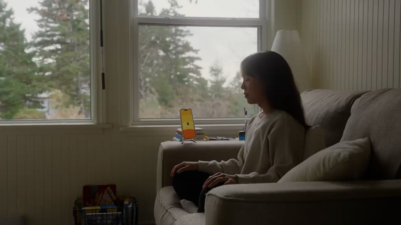 a young woman sits in profile on a couch in a living room bathed in soft natural light from a window. she is looking at a smartphone propped up on the armrest, possibly using a meditation app. the overall ambiance is calm and serene. the living room is cozy and inviting with muted, warm tones.