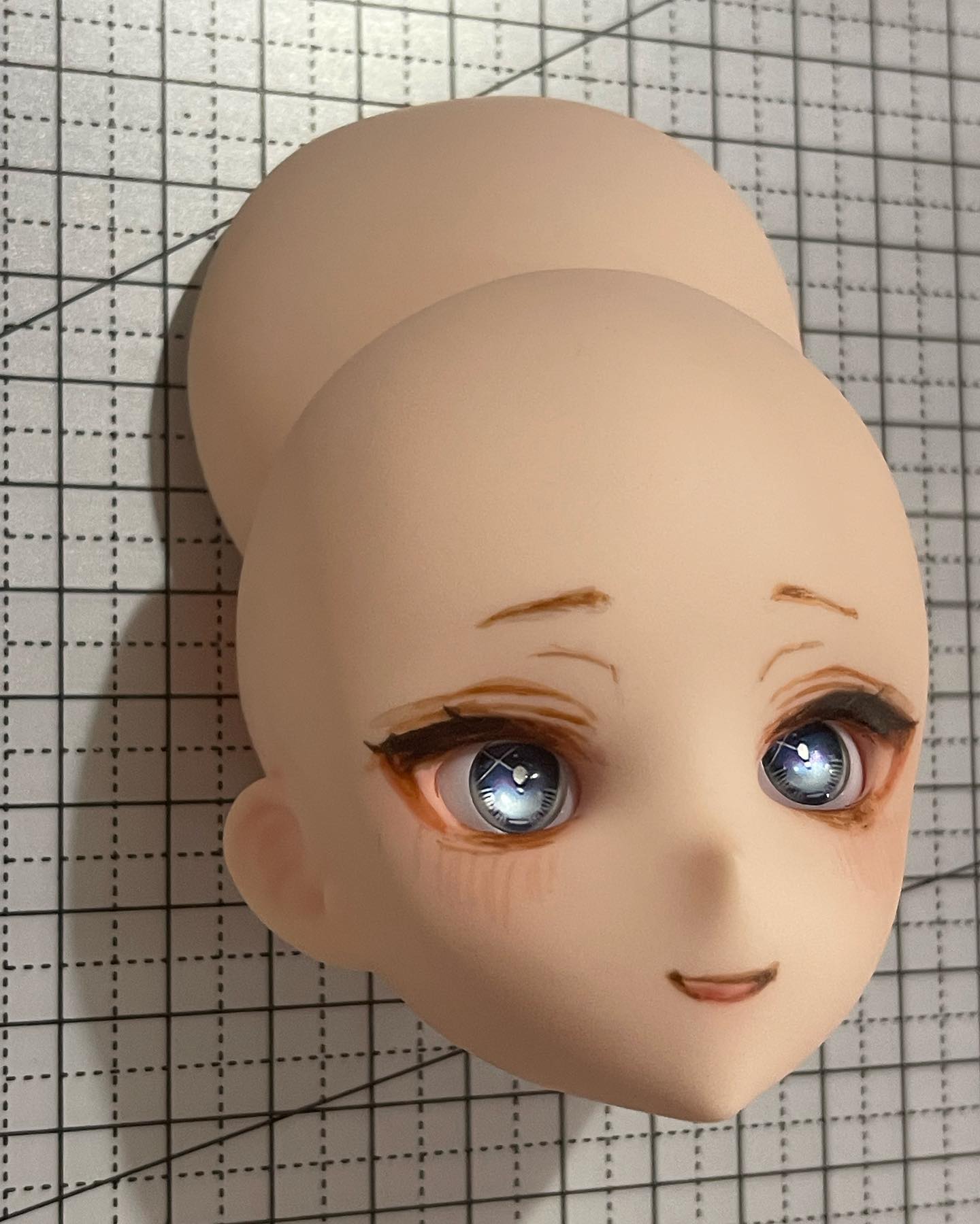 the image shows a closeup of a balljointed doll head. the head is made of a pale resin and has been painted with delicate features. the doll has large, blue eyes with a hint of sparkle, and its lips are slightly parted in a gentle smile. the doll's hair is styled in a bun, revealing the back of its head. the background of the image is a gray grid, which likely represents a crafting mat. the lighting in the image is artificial and highlights the doll's features. the perspective of the image is eyelevel, giving the viewer a direct look at the doll's face.