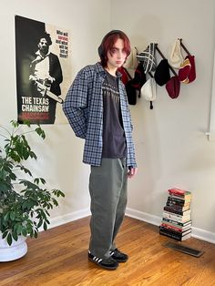 a man wearing a plaid shirt and green pants is standing in a room with a plant in a pot, a stack of books, and a wall with posters. he is looking off to the side.