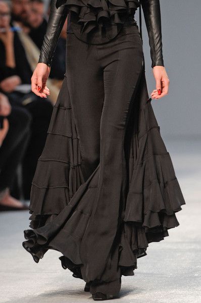 a woman with long black hair is wearing a black outfit on the runway with a skirt and pants that have ruffles. she is looking down, and her long black hair is flowing. she is walking with her head down, and her hands are at her side. her face is not visible. her outfit is very elegant, and she is wearing high heels.