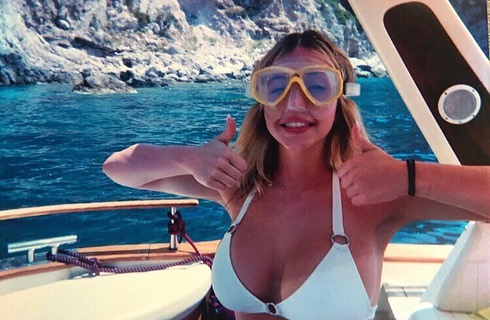 a young caucasian woman wearing a white bikini and yellow snorkeling goggles gives two thumbs up while on a boat in the mediterranean sea. the backdrop features clear blue water and rocky cliffs, suggesting a sunny summer vacation.  the woman appears happy and relaxed, enjoying her time on the boat. the photo seems to be taken from slightly above her level, showing the boat's details and the sea in the background. the overall impression is one of a carefree and enjoyable summer holiday.