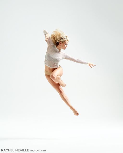 a blonde female dancer wearing a white longsleeved shirt and nude underwear is captured midleap against a white studio backdrop. the lighting is bright and even, highlighting the dancer's form and the graceful lines of her body. the overall aesthetic is minimal and elegant.