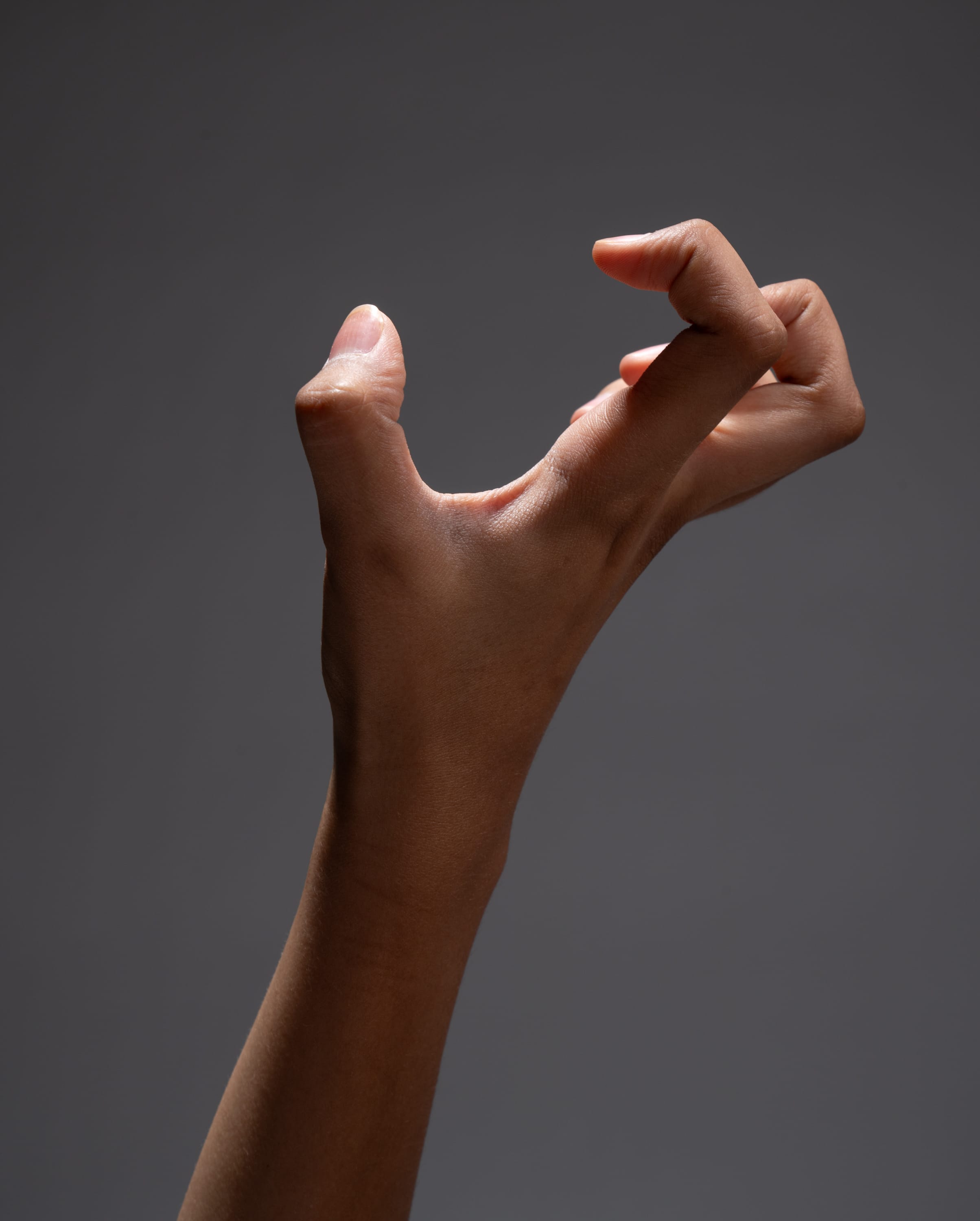 the image shows a hand reaching out to grab something with the index finger and thumb slightly touching. the fingers are curled in, while the hand is extended forward. the hand is illuminated with studio lighting, highlighting the details of the skin and the tendons. the background is a solid gray, which draws attention to the hand.