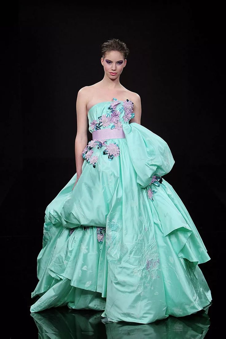 a model stands on a runway against a black background, wearing an aqua strapless taffeta gown with lilac floral embellishments and a lilac sash. the dress features a large, draped and voluminous skirt with cascading layers. the model has a light complexion and short, styled hair. the floor has a reflective surface.