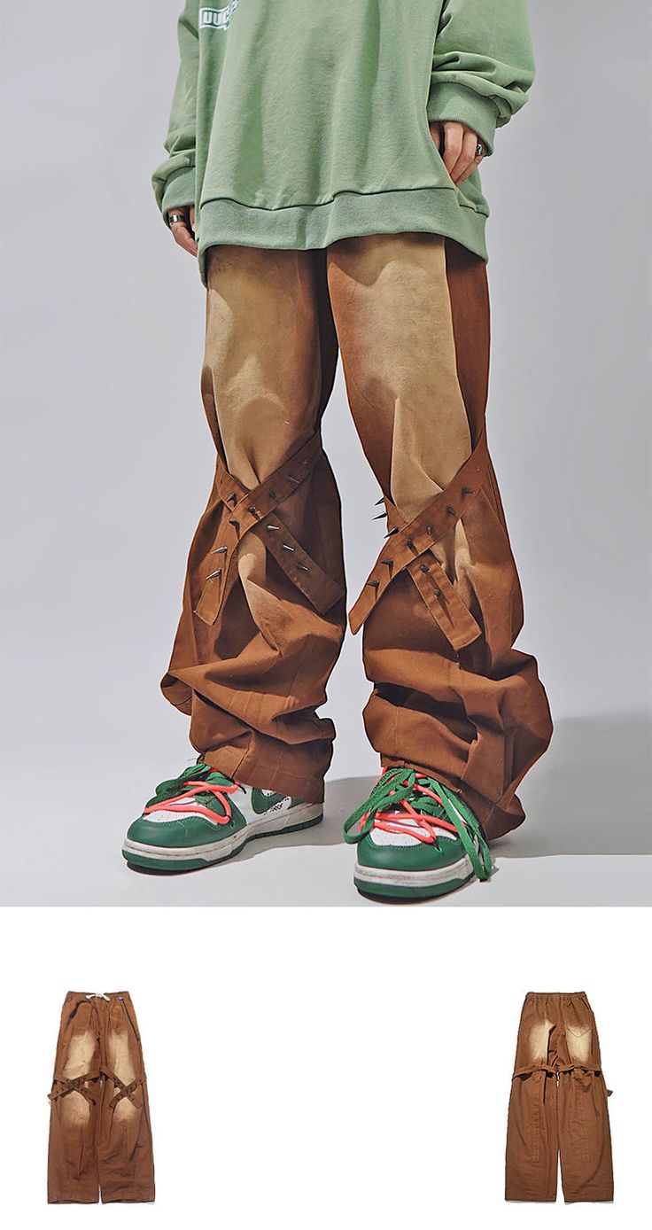 a man is wearing brown pants with a green sweatshirt and green and white sneakers. the pants are styled with unique brown straps across the front of the legs. the model is standing with his feet slightly apart.