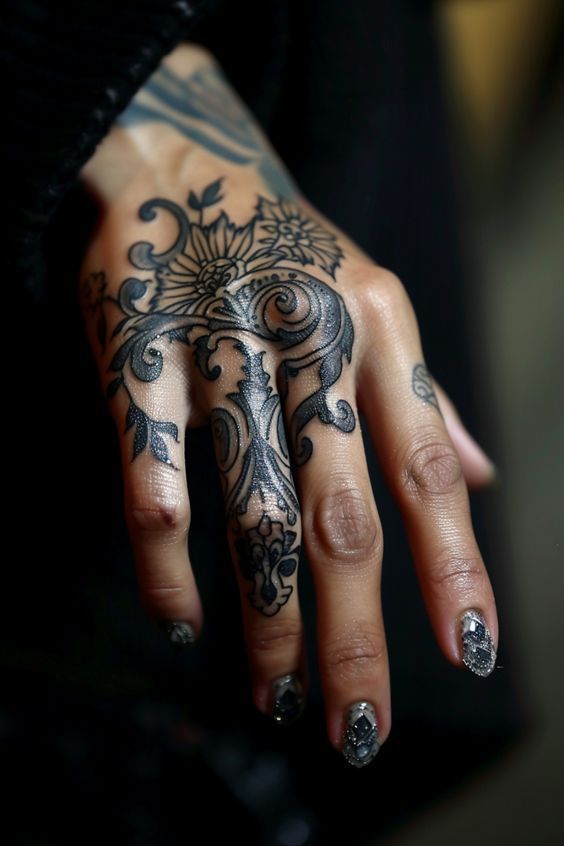 a closeup shot shows a person's hand adorned with an intricate black and white tattoo design, featuring flowers and ornamental patterns. the tattoo covers a significant portion of the hand and extends onto the middle finger. the person also sports sparkly nail polish on their nails.