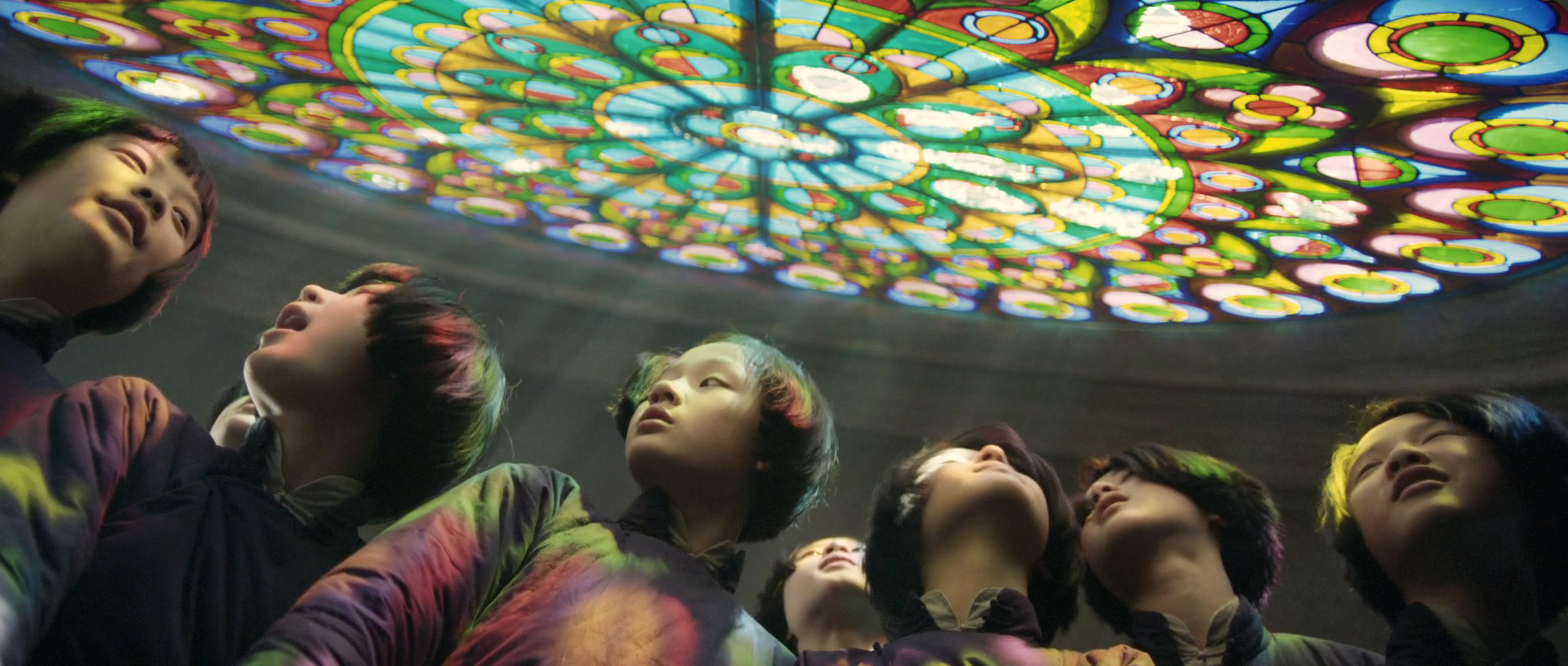 a group of children with colorful hair stand in a circle, their faces turned upwards in wonder.  a large, circular stained glass artwork is projected onto the ceiling above them, casting vibrant hues of green, blue, yellow, and red across their faces and clothing. the low angle shot emphasizes the children's perspective and the grandeur of the projected artwork.