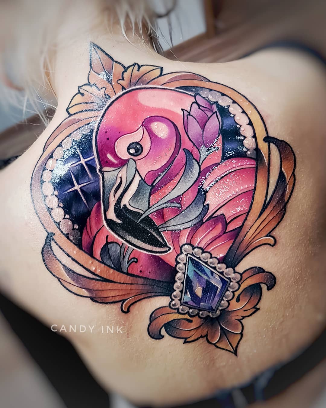 the image shows a woman's upper back with a fresh, colorful tattoo of a flamingo in a heartshaped, jewelencrusted frame. the flamingo is predominantly pink with purple accents, and the frame is primarily gold with a large purple jewel at the bottom. the tattoo is detailed and realistically rendered in a neotraditional style. the photo appears to have been taken shortly after the tattoo was completed, as the skin is still slightly red.