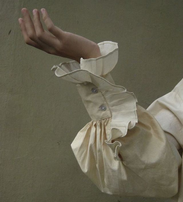 an arm clad in a creamcolored shirt with a ruffled sleeve is raised. the hand is positioned with the palm facing upward. the sleeve is detailed with ruffled edges and secured with two visible buttons.