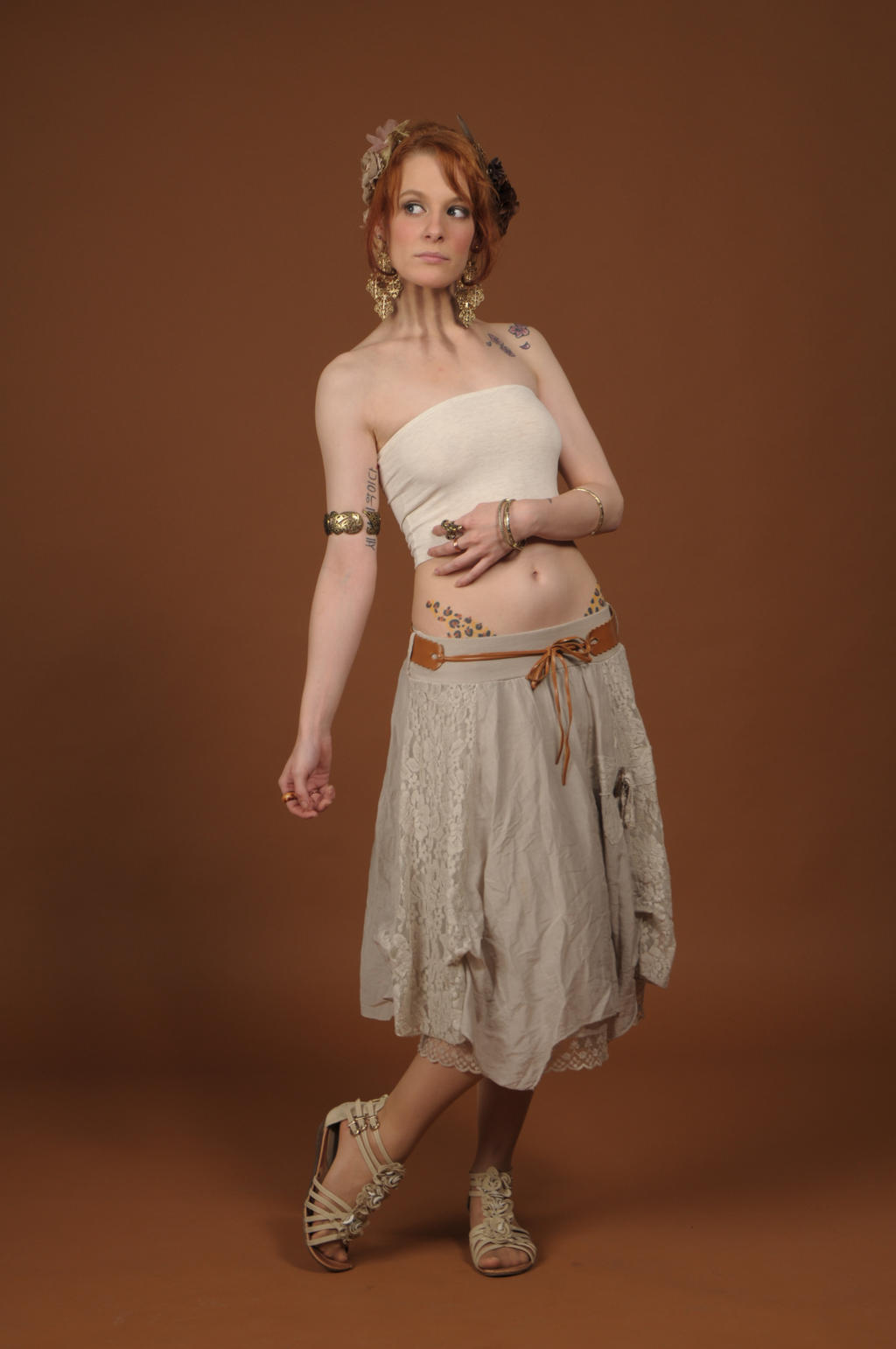a fulllength studio shot of a young redhead woman wearing a bohemianstyle outfit. she's wearing a light beige lace skirt, a white strapless top, brown sandals, several bracelets and earrings, and a floral hairpiece. a hennalike temporary tattoo is visible on her lower abdomen. the background is a solid brown.  the model is posed in a threequarter view, gazing slightly off to her right.