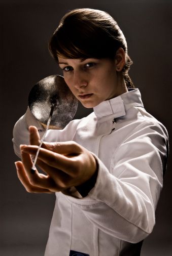 a woman in a white fencing uniform stands indoors with a foil, her hand on the grip, her face shows determination and focus. the image is a close up, the background is dark and blurry.