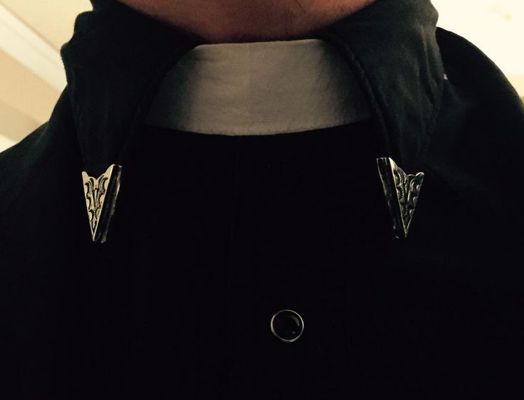 a closeup shot of a man wearing a black shirt with a white clerical collar. two silver collar pins are attached to the collar, and one button is visible on the shirt.