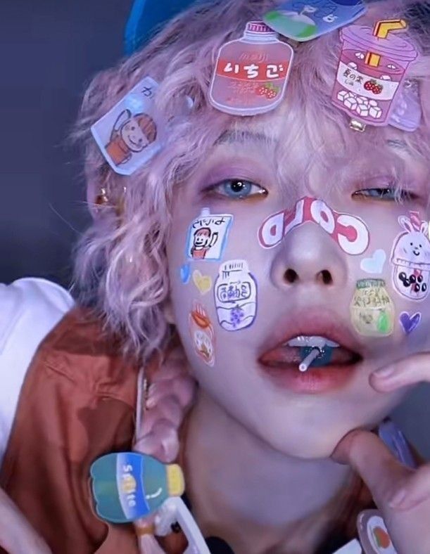 a closeup selfie of a young person with dyed pink hair, their face decorated with various playful stickers, primarily featuring japanese text and cartoon imagery. the individual has bluecolored contact lenses and is