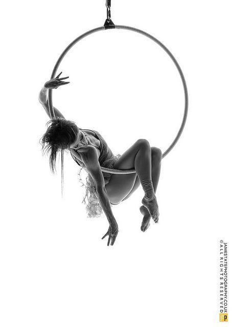 a black and white photograph of a woman in a hoop. the woman is suspended upside down and hanging from the hoop. she is bending her body in a unique pose.  the woman is wearing a black sports bra. the photograph is set against a white background.