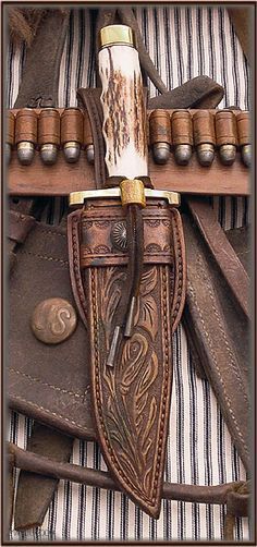 a closeup of a westernstyle knife with a staghorn handle, leather sheath with tooled design, and an ammunition belt, all arranged on a striped cloth background. the ensemble suggests a historical or vintage theme.